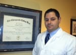 A man in white lab coat standing next to a framed certificate.